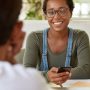 Content glad woman with Afro haircut, wears piercing, holds smart phone, sits opposite her fellow, c